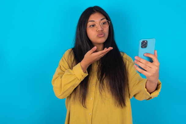 mladá latinská žena nosí žlutou košili přes modré pozadí fouká vzdušný polibek na kameru smartphone a bere selfie, posílá mwah přes on-line volání. - Fotografie, Obrázek