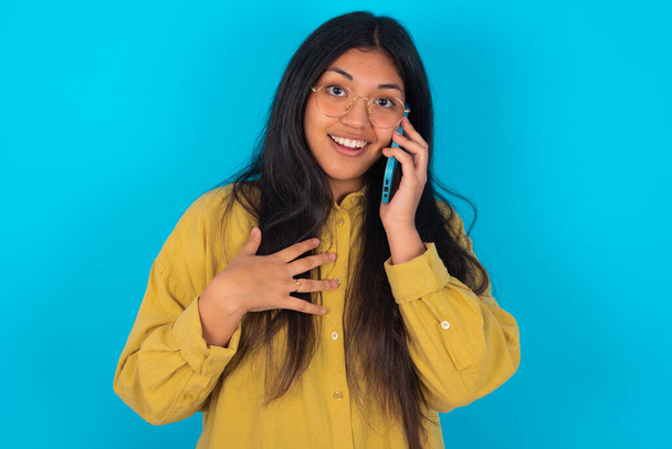 Smiling young latin woman wearing yellow shirt over blue background talks via cellphone, enjoys pleasant great conversation. People, technology, communication concept - Photo, Image