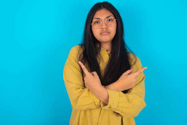 Jovem mulher latina séria vestindo camisa amarela sobre fundo azul cruza mãos e pontos em lados diferentes hesita entre dois itens. Conceito de decisão difícil - Foto, Imagem