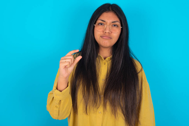Mavi arka planda sarı gömlek giyen hoşnutsuz genç Latin kadın küçük el işaretleri çok büyük olmayan bir şeyi gösteriyor. Vücut dili kavramı. - Fotoğraf, Görsel