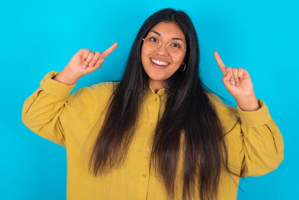 Cheerful young latin woman wearing yellow shirt over blue background demonstrating hairdo - Фото, изображение