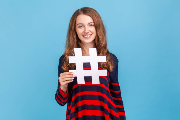 Attractive woman wearing striped casual style sweater, holding white hashtag symbol, tagged message, popular internet idea, viral content. Indoor studio shot isolated on blue background. - Foto, Bild