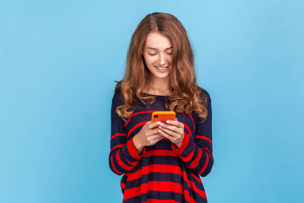 Happy woman wearing striped casual style sweater, with pleasant smile using cell phone, answering messages in social networks, writing post. Indoor studio shot isolated on blue background. - Fotoğraf, Görsel