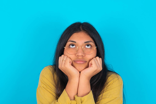 Portrait of sad young latin woman wearing yellow shirt over blue background hands face look empty space - Foto, imagen