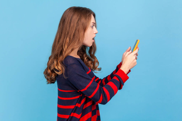 Side view of shocked woman in striped casual style sweater, holding smart phone in hands, looking at display with astonished expression, braking news. Indoor studio shot isolated on blue background. - Fotografie, Obrázek