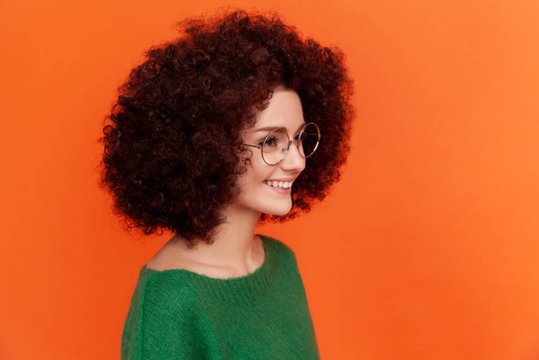 Side view portrait of attractive woman with Afro hairstyle with perfect skin wearing green casual style sweater and spectacles, looking away, smiling. Indoor studio shot isolated on orange background. - Photo, image
