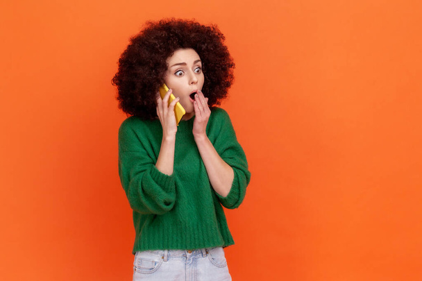 Ohromená žena s afro účesem v zeleném neformálním stylu svetru mluví přes mobil, slyší šokující zprávy, zakrývá ústa dlaní. Vnitřní studio záběr izolované na oranžovém pozadí. - Fotografie, Obrázek