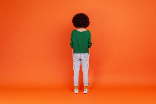 Back view of slim woman with Afro hairstyle wearing green casual style sweater and jeans standing and looking at something, waiting. Indoor studio shot isolated on orange background. - Фото, изображение