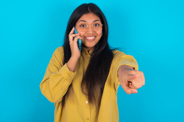 Positivo giovane donna latina che indossa camicia gialla su sfondo blu indica direttamente alla fotocamera ha conversazione telefonica sorride ampiamente gode di parlare lunghe ore. Unisciti a me. - Foto, immagini