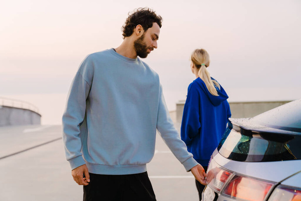 Young man and woman standing together by car on parking outdoors - Photo, Image