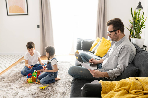 Busy single parent is babysitting and working on the distance. Father using laptop for freelance work sitting on the sofa at home while his two kids playing peacefully together - 写真・画像