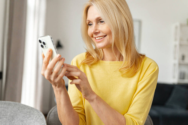 Glad mature woman talking online on mobile phone via video connection, participating in a conference, sitting at the table video-calling - 写真・画像