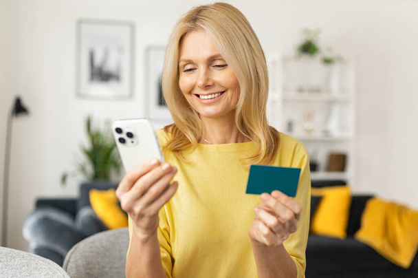 Satisfied smiling mature woman sitting on the couch, holding credit card and smartphone, paying in the Internet, making order, charming lady enjoys shopping - Foto, immagini