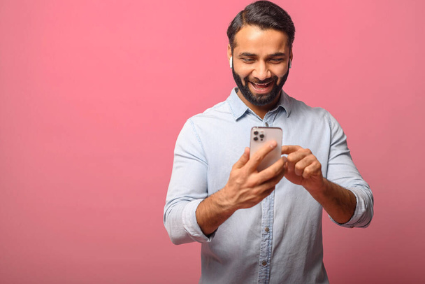 Smiling Indian man holding and using smartphone, carefree multiracial guy enjoying online chatting - 写真・画像