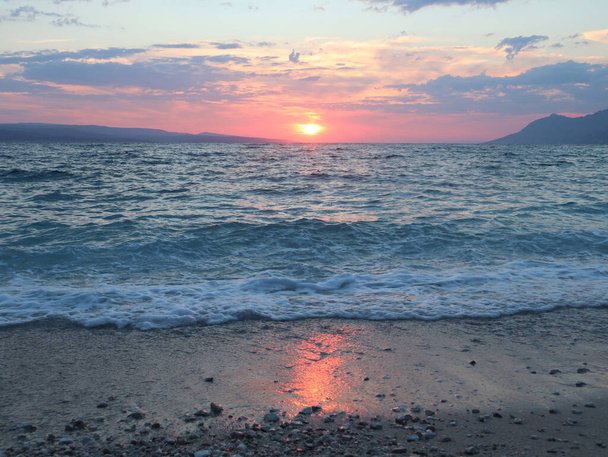 Merimaisema merenrannalla auringonlaskun aikaan kesäiltana. Auringonlasku yli meren, taivas ja hiekka rannalla valaisevat auringon säteet vaaleanpunainen, Kroatia, Dalmatia, Brela - Valokuva, kuva