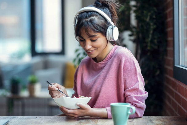 Shot of beautiful young woman listening music with headphones while having healthy breakfast in the living room at home. - Foto, Imagem