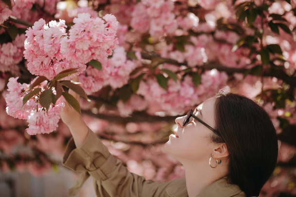 Happy young asian woman standing under the branches of sakura and admiring the flowering branches. Lady looking at pink blooming flovers  - Fotoğraf, Görsel