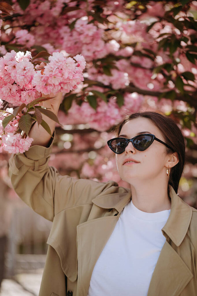 Fashion woman in black sunglasses posing on the camera under blooming sacura tree branches - Valokuva, kuva