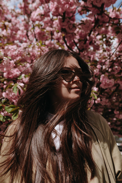 Portré szépség nő remegő hosszú barna haj napszemüvegben a virágos fa háttér - Fotó, kép