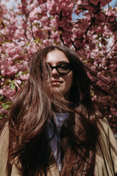 Portrait de jolie femme aux longs cheveux bruns volants en lunettes de soleil sur le fond d'arbre fleuri - Photo, image
