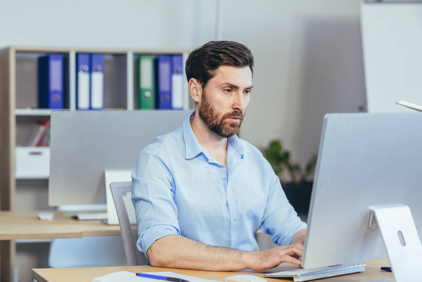 Portrait of a serious and focused businessman, a man working in a bright office during the day, at the computer looking at the monitor, in casual clothes - Photo, Image