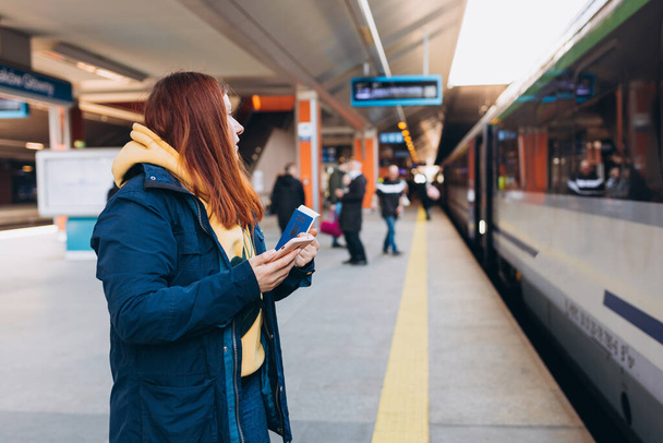 Nuori punapää nainen odottaa asemalla laiturilla reppu taustalla sähköinen juna älypuhelimella. Rautatieliikenteen konsepti, Matkustaja. Hyökkäys Ukrainassa. - Valokuva, kuva