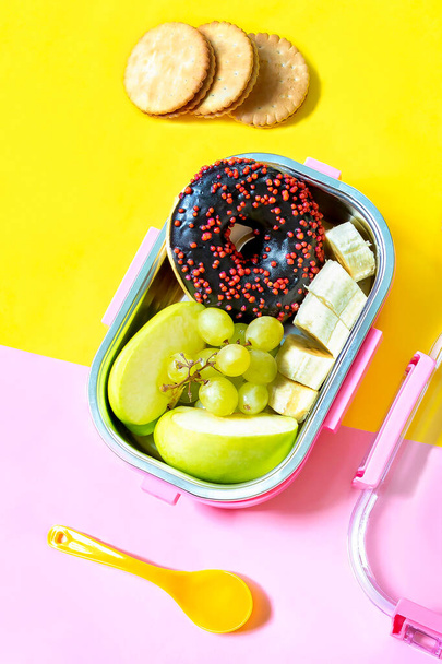 Catering food prepared in storage container with compartment with doughnut,apple,banana,cookies on pink background. meal with healthy balanced diet, lunch box boxed take away delivery packed ready. - Fotó, kép