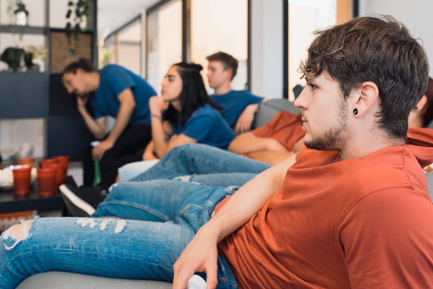 группа молодых людей, скучающих, смотрящих футбол по телевизору. уставшие юные друзья, на домашней вечеринке. отдых - Фото, изображение