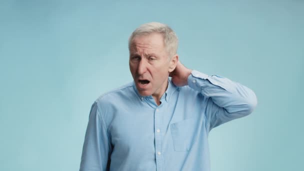 Problemas de salud del envejecimiento. trastornado hombre mayor que sufre de dolor de cuello, calentamiento y masaje parte del cuerpo, fondo azul - Imágenes, Vídeo