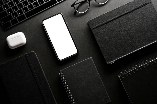 Modern black office workspace background with office accessories and devices mockup. Smartphone white screen mockup, notebook, earbuds, eyeglasses and objects - Photo, Image