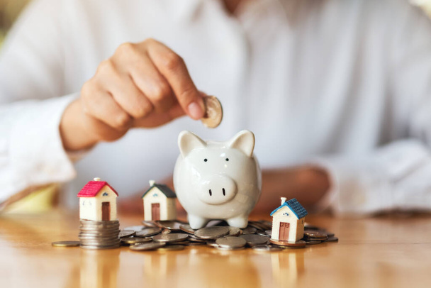 A woman putting coins into piggy bank with house models and pile of coins on the table for saving money concept - Photo, image