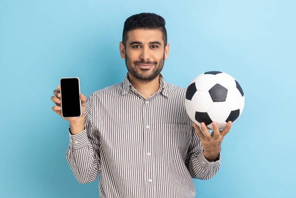 Smiling satisfied bearded man holding soccer ball and smartphone empty black display, ticket booking for championship, wearing striped shirt. Indoor studio shot isolated on blue background. - Photo, image