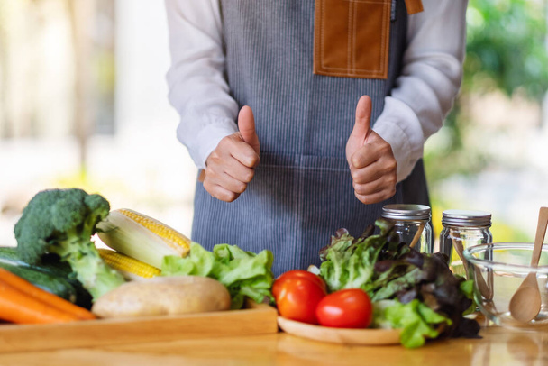 Nahaufnahme einer Köchin, die Handzeichen macht und Daumen nach oben zeigt, während sie frisches gemischtes Gemüse zubereitet, um in der Küche zu kochen - Foto, Bild