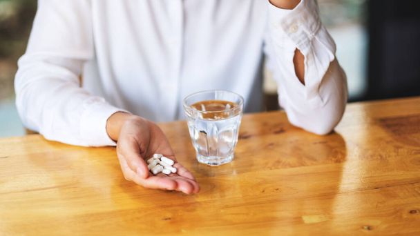 Closeup image of a woman holding white pills with a glass of water on the table - Photo, Image