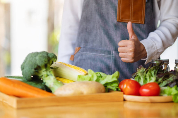 Immagine ravvicinata di una cuoca che fa e mostra i pollici in alto segno della mano mentre prepara verdure fresche miste per cucinare in cucina - Foto, immagini