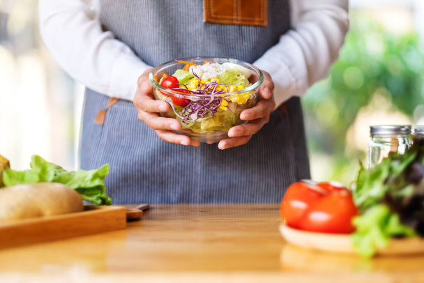 Immagine ravvicinata di una cuoca che cucina e tiene una ciotola di insalata di verdure miste fresche in cucina - Foto, immagini