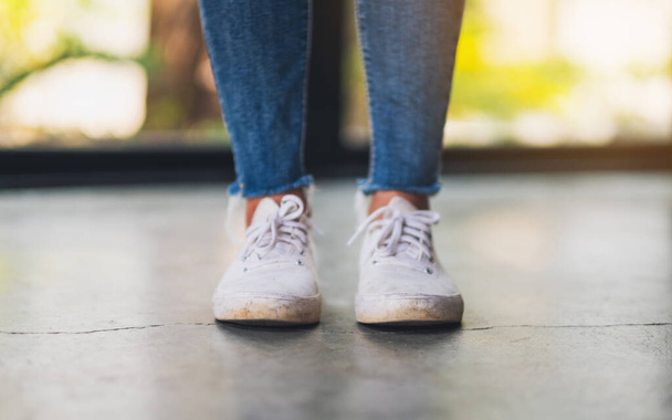 Closeup image of a woman wearing jean and white sneakers standing on concrete floor - Photo, image