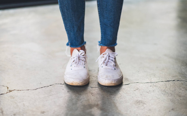Imagem de close-up de uma mulher vestindo jeans e tênis brancos em pé no chão de concreto - Foto, Imagem