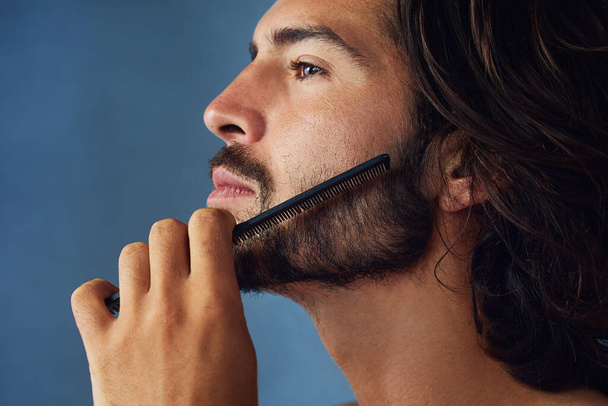 Taming his mane. Studio shot of a handsome young man grooming his facial hair against a blue background. - Foto, Imagen