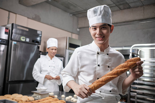 Beyaz üniformalı genç Asyalı aşçı portresi kamera, mutlu bir gülümseme ve baget ekmeği ile neşeli, profesyonel yemek işi, bir restoran mutfağında ticari hamur işi. - Fotoğraf, Görsel