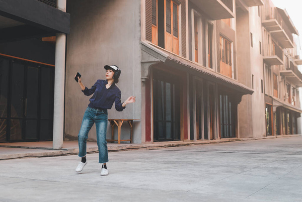 Szczęśliwa młoda azjatycka kobieta słuchająca muzyki i bawiąca się ze słuchawkami za pośrednictwem smartfona na ulicy. - Zdjęcie, obraz
