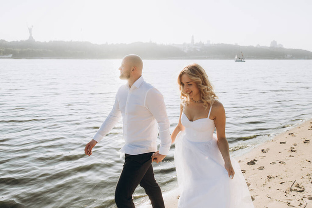 Esküvői fotózás napnyugtakor egy gyönyörű pár, a menyasszony fehér ruhában, és a vőlegény fehér ingben és fekete nadrágban - Fotó, kép