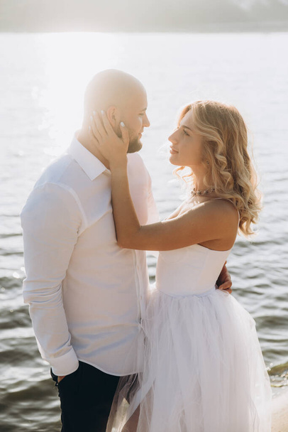 Fotosesión de boda al atardecer de una hermosa pareja, la novia con un vestido blanco, y el novio con una camisa blanca y pantalones negros - Foto, imagen