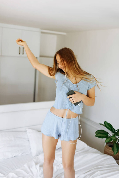 Young teen girl in pajama standing on her bed and singing a song. Cheerful caucasian adolescent girl with headphones dancing on her bedroom  - Fotó, kép
