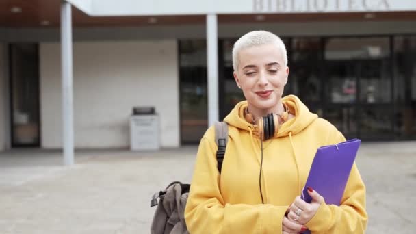 Estudiante universitaria con el pelo rubio corto afeitado Sonriendo mirando a la carpeta de la cámara fuera del edificio de la universidad - Metraje, vídeo