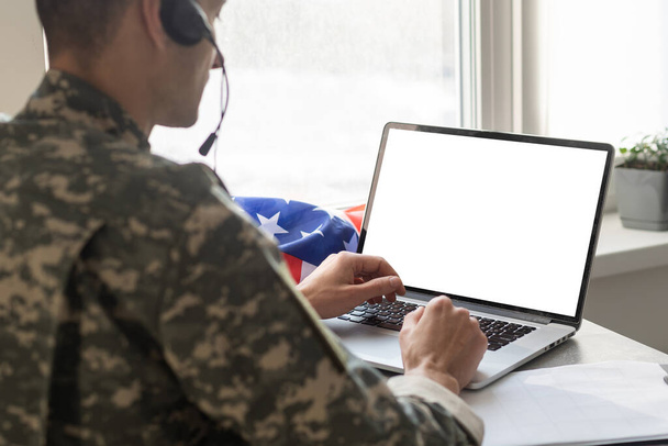 Στρατιωτικός άνθρωπος gesturing στο γραφείο κοντά στο φορητό υπολογιστή με λευκή οθόνη, σημαία ΗΠΑ. - Φωτογραφία, εικόνα