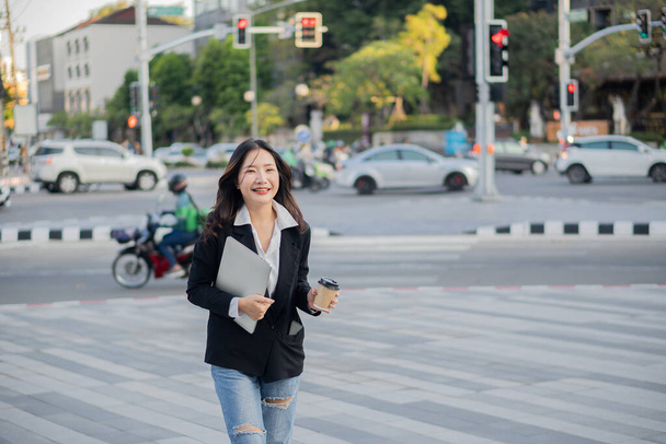 Una joven mujer de negocios asiática con un portátil y una taza de café entra en la oficina felizmente. - Foto, imagen