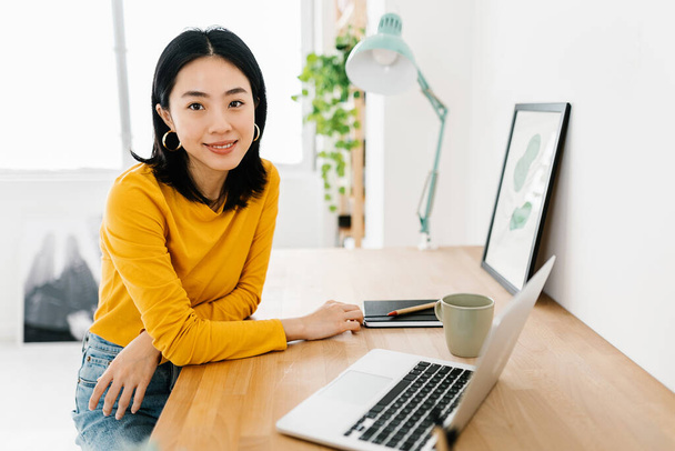 Portrait of a young asian woman looking at camera while sitting on work desktop - Valokuva, kuva