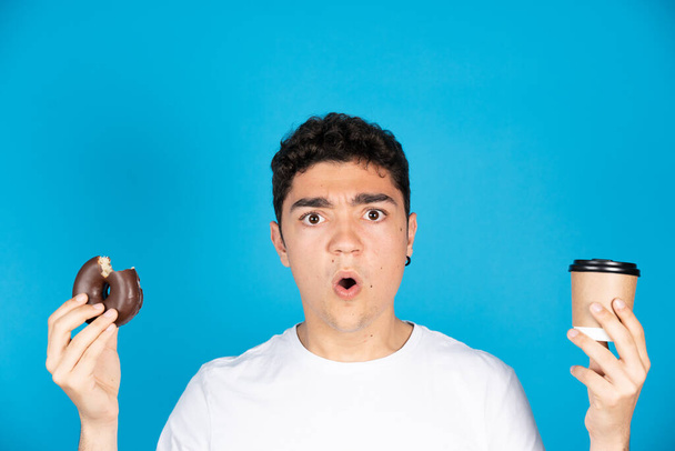 Surprised hispanic teenager holding a disposable cup of coffee and a donut or doughnut isolated on blue background - Photo, image