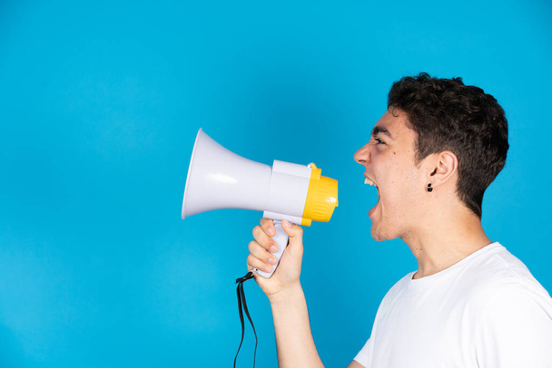 Hispanic teenager boy shouting on megaphone or speaker to copy space isolated on blue background. - Foto, Imagem